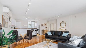A modern living room with a black leather sofa, a glass coffee table, and a dining area with a white kitchen in the background.