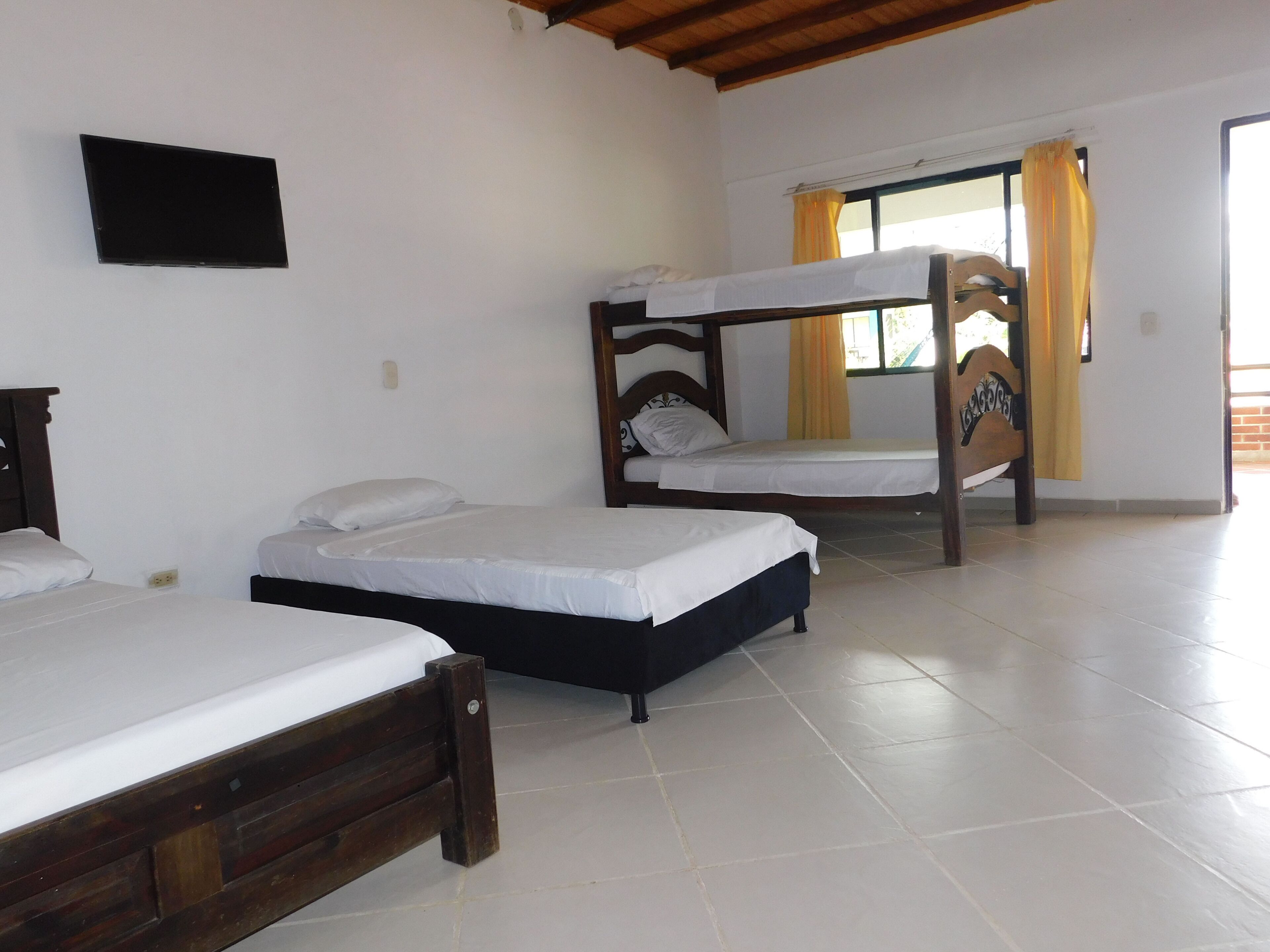 A room with three wooden beds, a flat-screen TV mounted on the wall, and a window with yellow curtains.