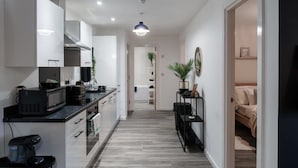 Una cucina moderna con mobili bianchi, un piano di lavoro nero e una macchina da caffè. Si apre un corridoio che conduce a una camera da letto con un letto e una pianta.