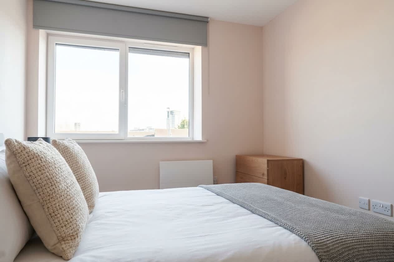 Et soveværelse med seng, to puder, en radiator, et trænatbord og et vindue.