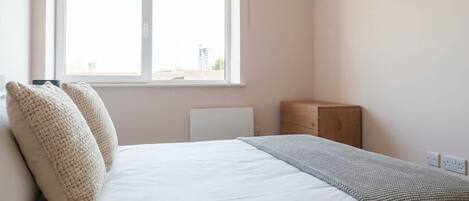 A bedroom with a bed, two pillows, a radiator, a wooden nightstand, and a window.