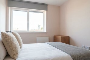 Une chambre à coucher avec un lit, deux oreillers, un radiateur, une table de chevet en bois et une fenêtre.
