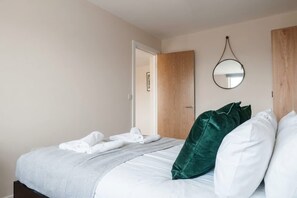A neatly made bed with white linens, a green pillow, and a round mirror on the wall.