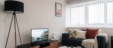A modern living room with a TV on a wooden cabinet, a floor lamp, a sofa with cushions, a coffee table with books and a plant, and a window with blinds.