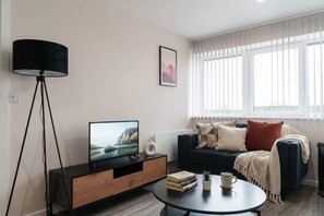 A modern living room with a TV on a wooden cabinet, a floor lamp, a sofa with cushions, a coffee table with books and a plant, and a window with blinds.