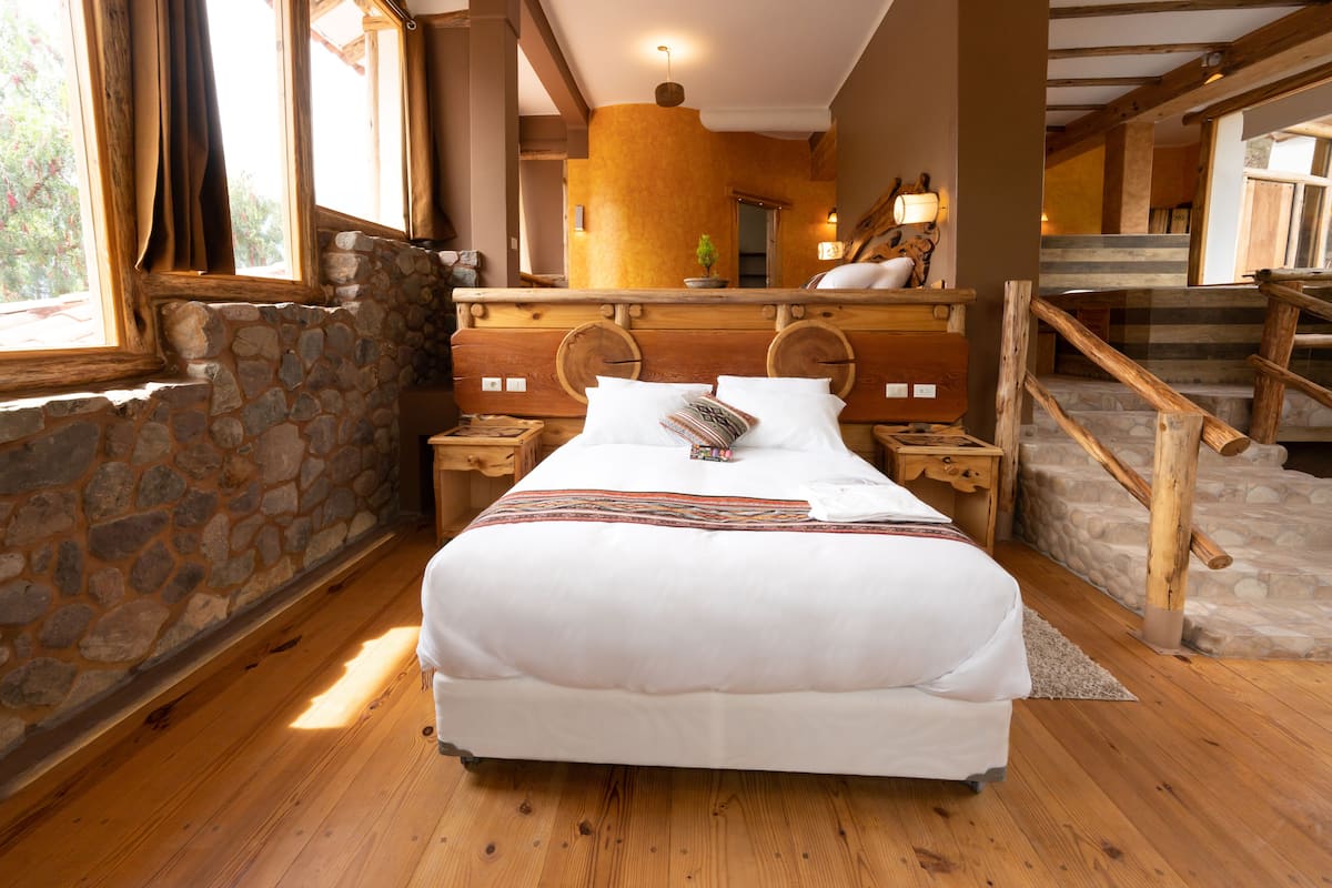 A wooden-framed bed with a stone wall and wooden floor, a wooden headboard, and a small nightstand with a lamp.