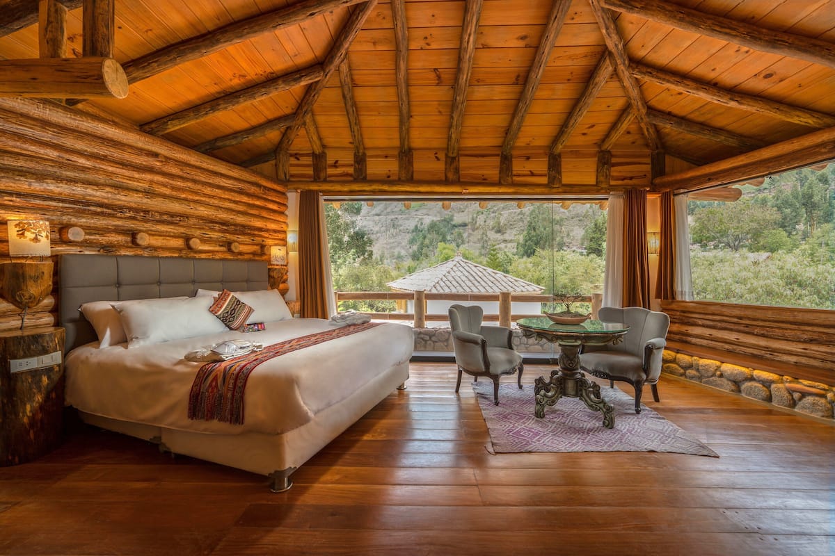 A log cabin bedroom with a large bed, a dining table with chairs, and a view of the surrounding forest.