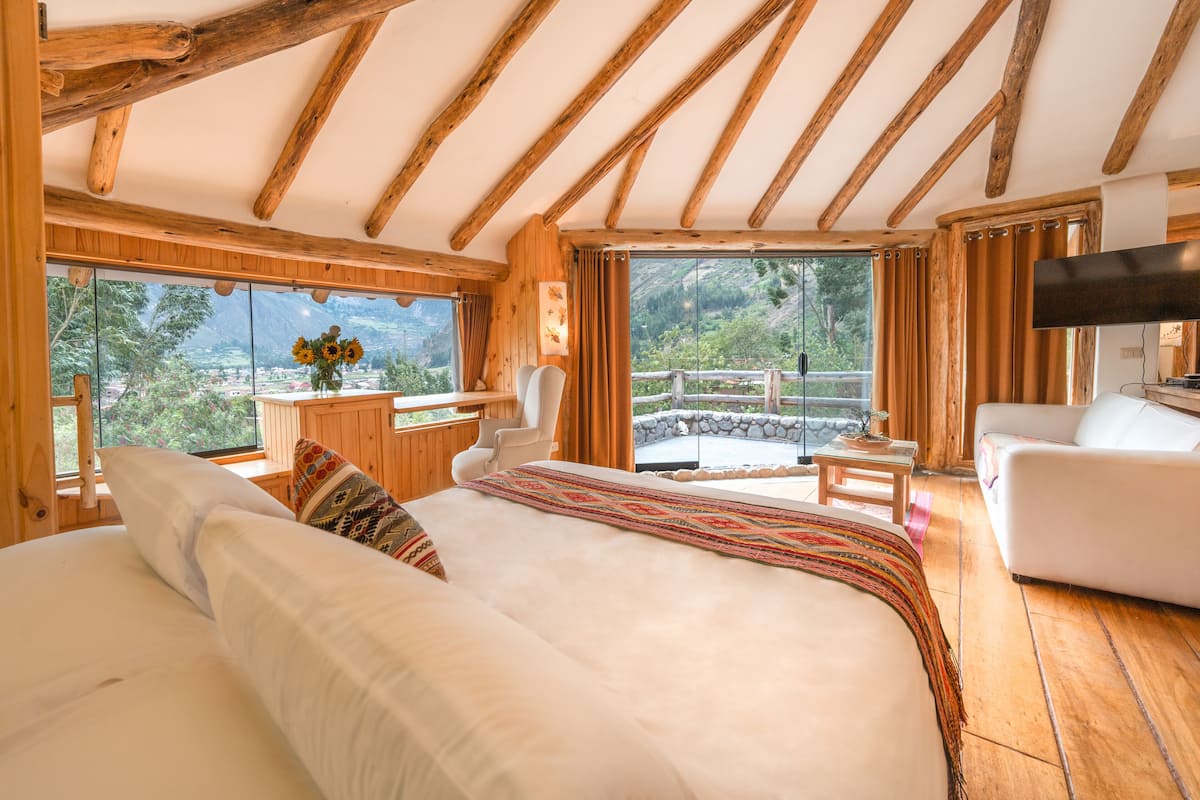 A bedroom with a large bed, a desk, a chair, and a TV. There is a balcony with a view of the outdoors.
