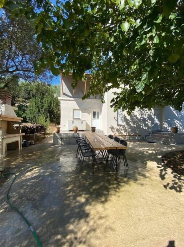 Un espacio de comedor al aire libre con una mesa y sillas de madera, rodeado de árboles y un edificio.