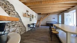 Un intérieur rustique avec un mur en pierre, des poutres en bois au plafond, un coin cuisine équipé d’une cuisinière et un espace repas avec des chaises et des tables.