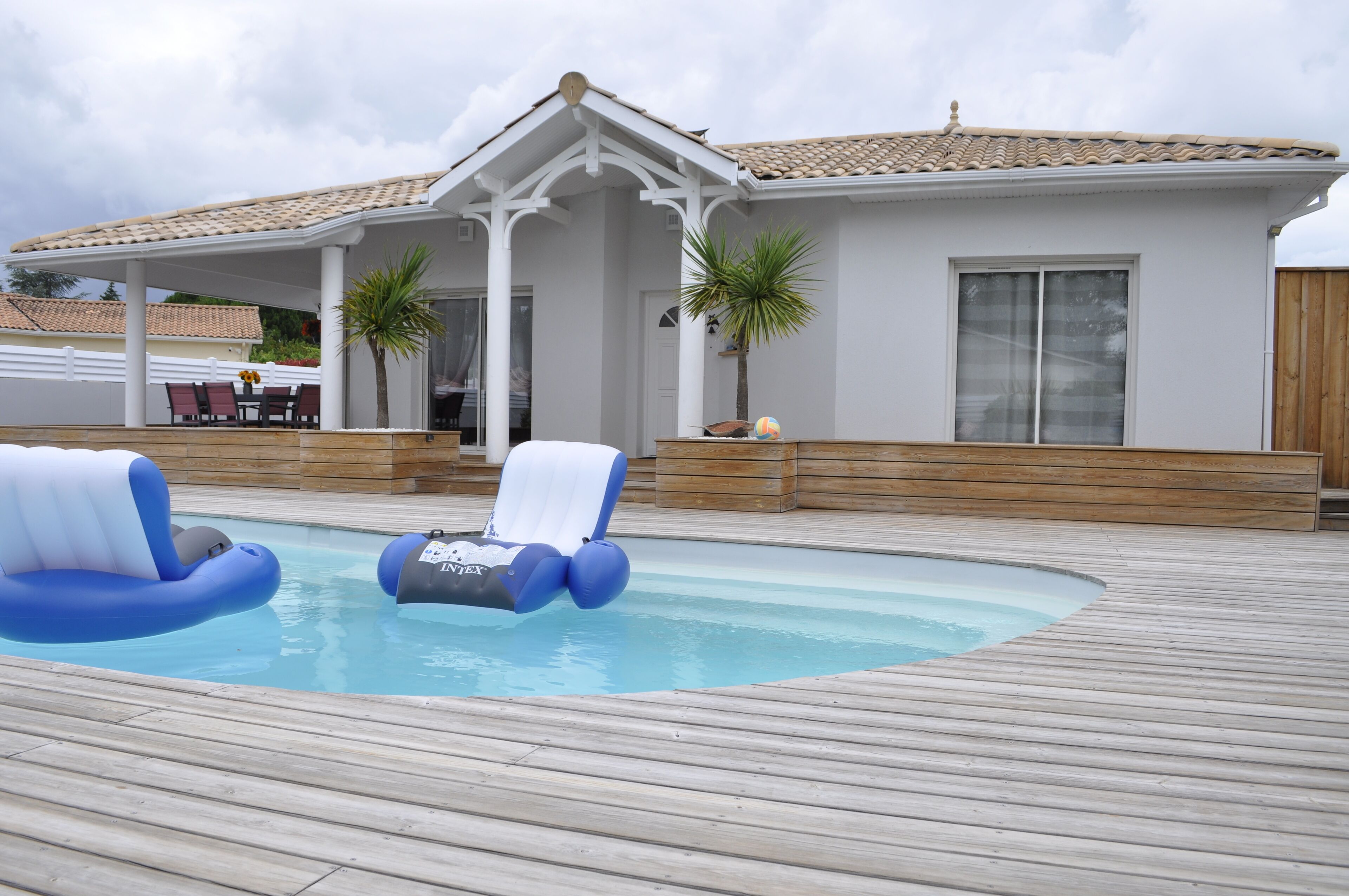 A swimming pool with two blue lounge chairs, a covered patio area, and a white building with a tiled roof.