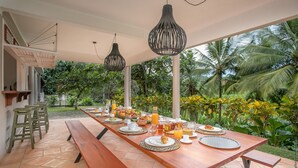 A long wooden dining table set with breakfast items, surrounded by green chairs and hanging pendant lights, with a view of lush tropical vegetation. - Habitation La Thibault Exceptional estate in the heart of Martinique (Gros-Morne)