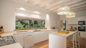 A modern kitchen with white cabinetry, a central island, a gas stove, and a large window with a view of tropical foliage. - Habitation La Thibault Exceptional estate in the heart of Martinique (Gros-Morne)