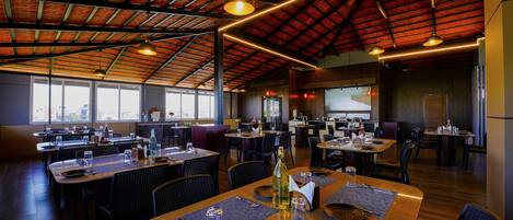A spacious restaurant with high ceilings, multiple dining tables, and a view of the city through large windows.