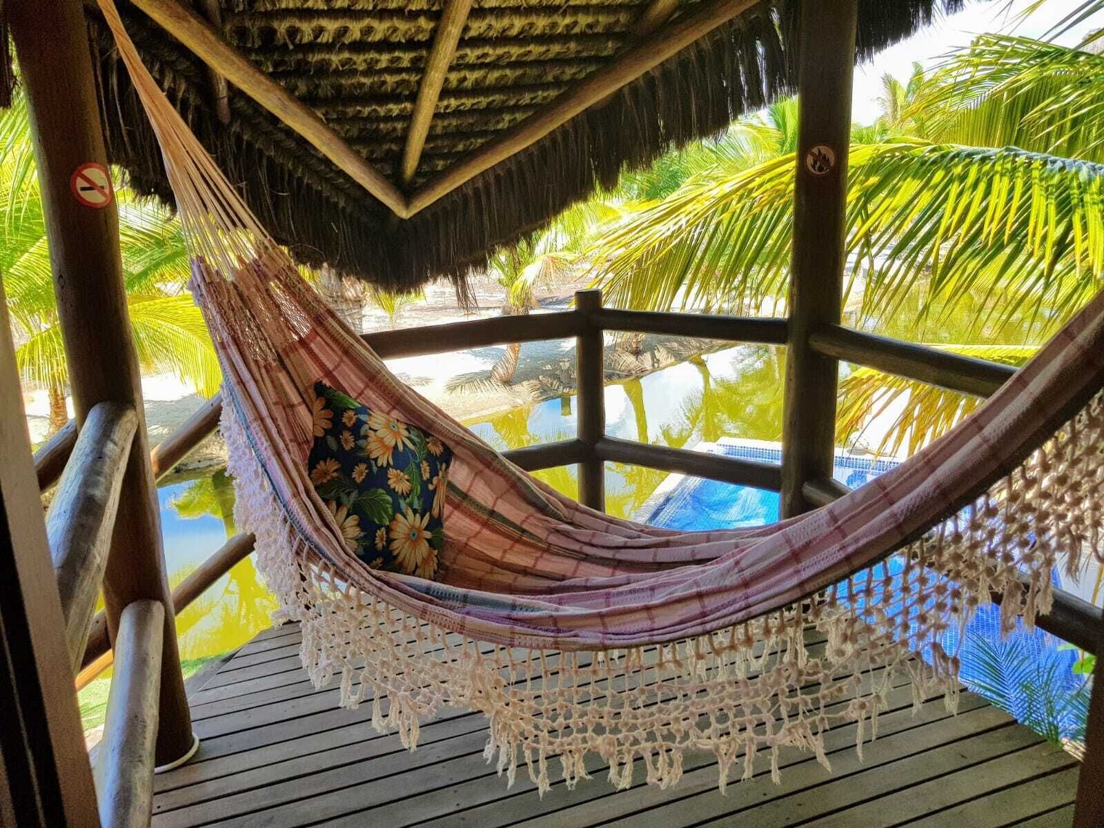 A hammock under a thatched roof, overlooking a pool and palm trees.