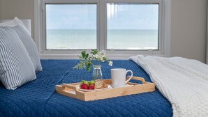 Una cama con un edredón azul, una almohada blanca, una bandeja con fresas, una taza y un florero con flores.