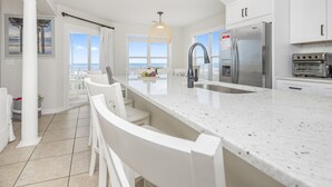 Una cocina moderna con armarios blancos, una isla grande con taburetes de bar, un refrigerador de acero inoxidable y vistas al océano a través de amplias ventanas.