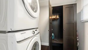 Une buanderie équipée d’un lave-linge et d’un sèche-linge Samsung, d’une porte en bois foncé et d’un petit espace de rangement où sont suspendus des vêtements.