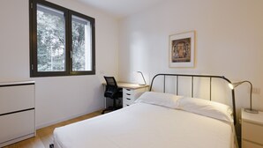 Un dormitorio con cama, escritorio, silla, mesita de noche y ventana con vista a árboles.