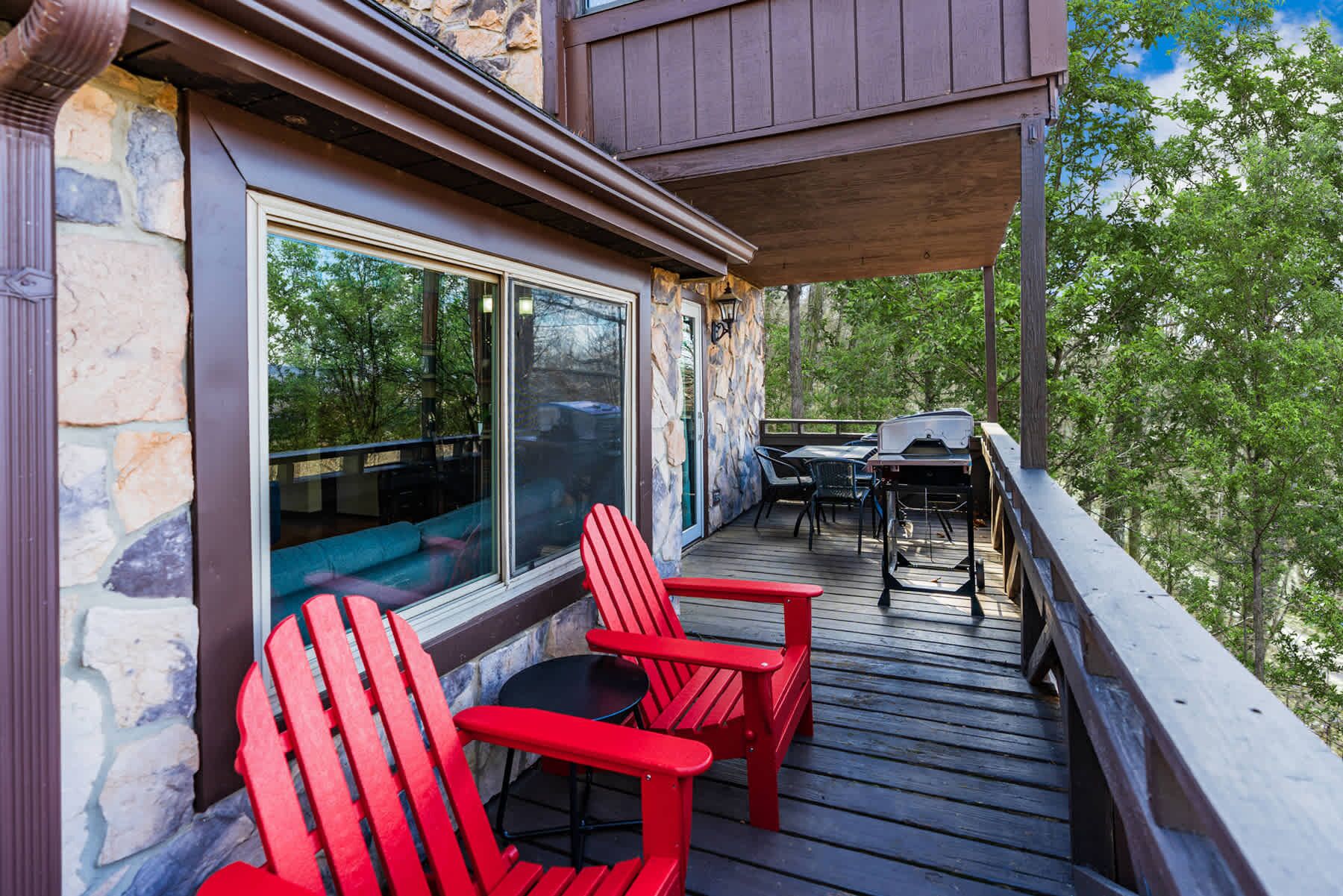 A wooden deck with red Adirondack chairs and a table, overlooking a forested area.