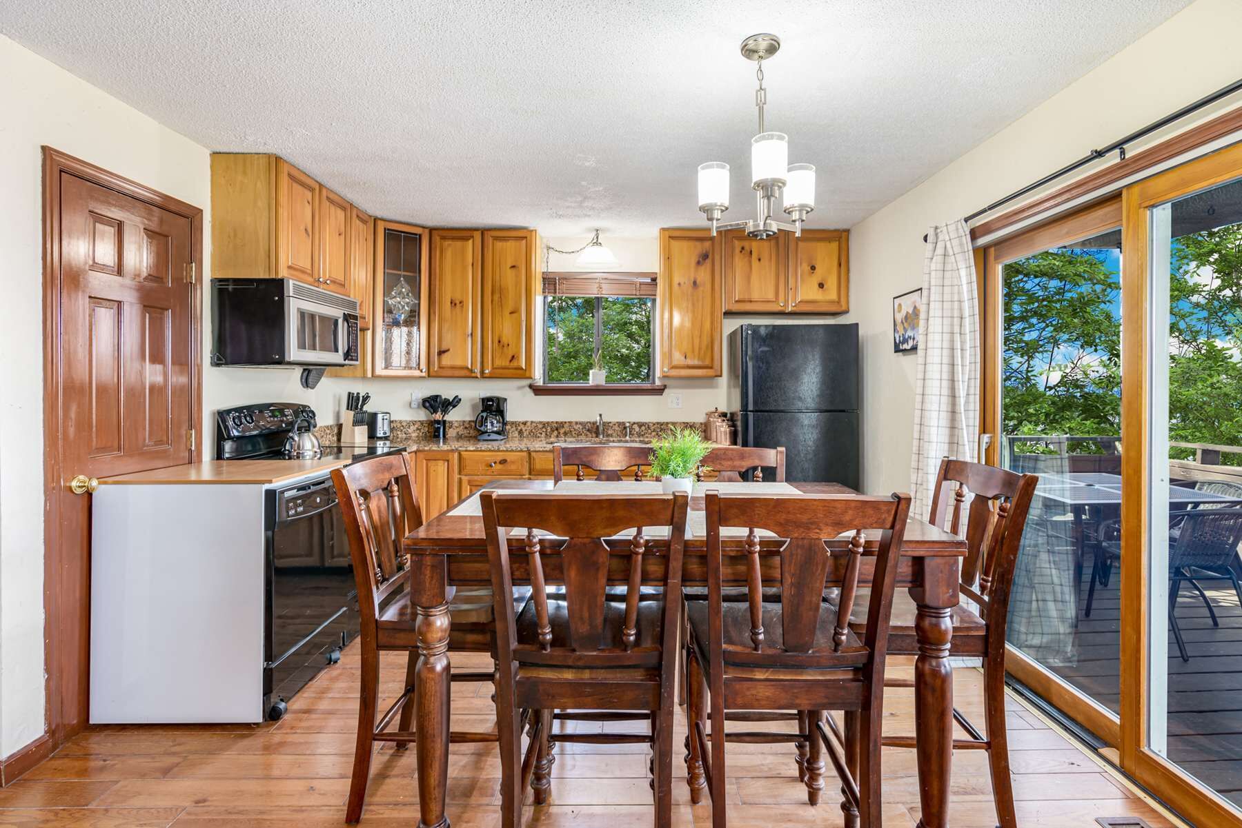 A kitchen with wooden cabinets, a dining table with chairs, a refrigerator, a microwave, and a window with a view of trees.