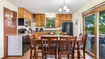 A kitchen with wooden cabinets, a dining table with chairs, a refrigerator, a microwave, and a window with a view of trees.