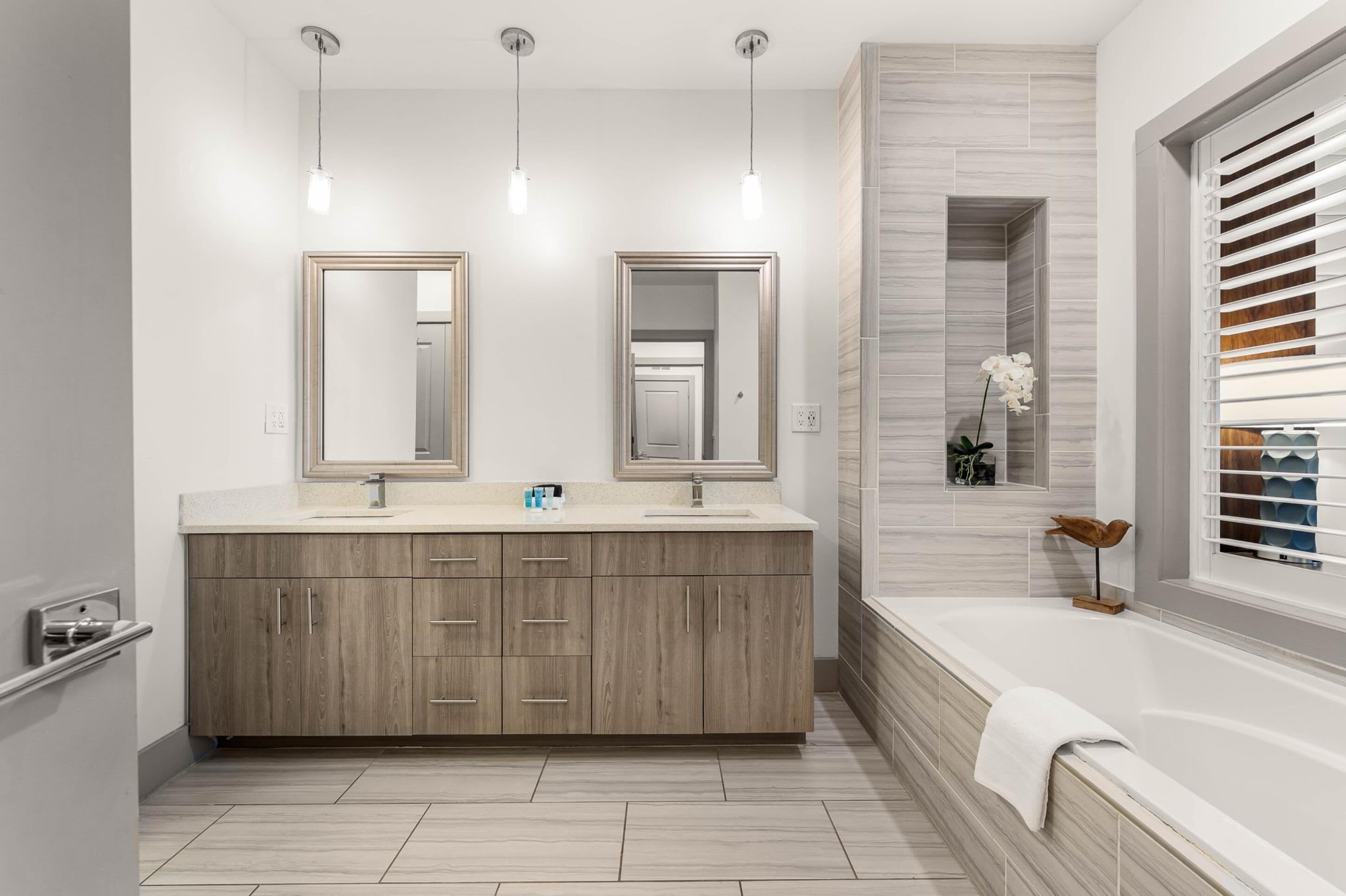 A modern bathroom with a wooden vanity, two mirrors, a large bathtub, and a window with blinds.