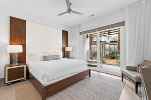 Ein modernes Schlafzimmer mit einem großen Bett, zwei Nachttischen mit Lampen, einem Deckenventilator und Blick auf einen Garten durch große Schiebetüren.