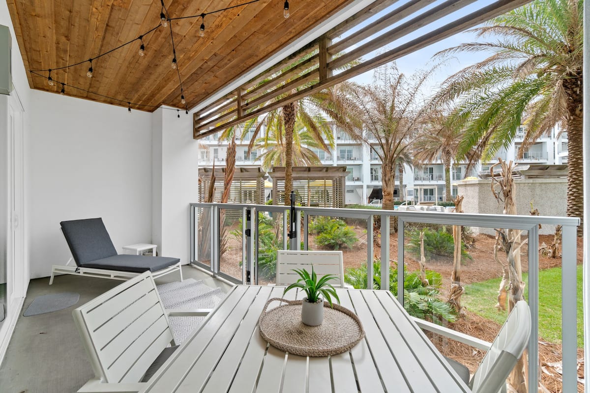 Een balkon met houten vloer, een witte tafel met geweven onderzetter, een lounge stoel en een kleine witte tafeltje.