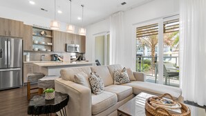 Een moderne keuken met een koelkast, magnetron en houten kastjes. Er staat een beige bank met decoratieve kussens en een klein tafeltje met een mandje.