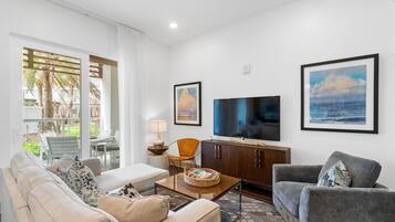 Una sala moderna con sofá, sillón, mesa de centro y televisor. Cuenta con un balcón con mobiliario exterior y palmeras.