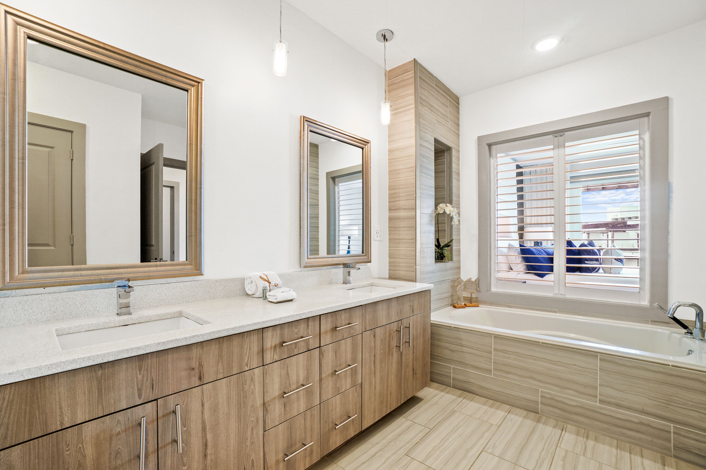 Un baño moderno con un tocador amplio, dos espejos, una bañera y una ventana con persianas.
