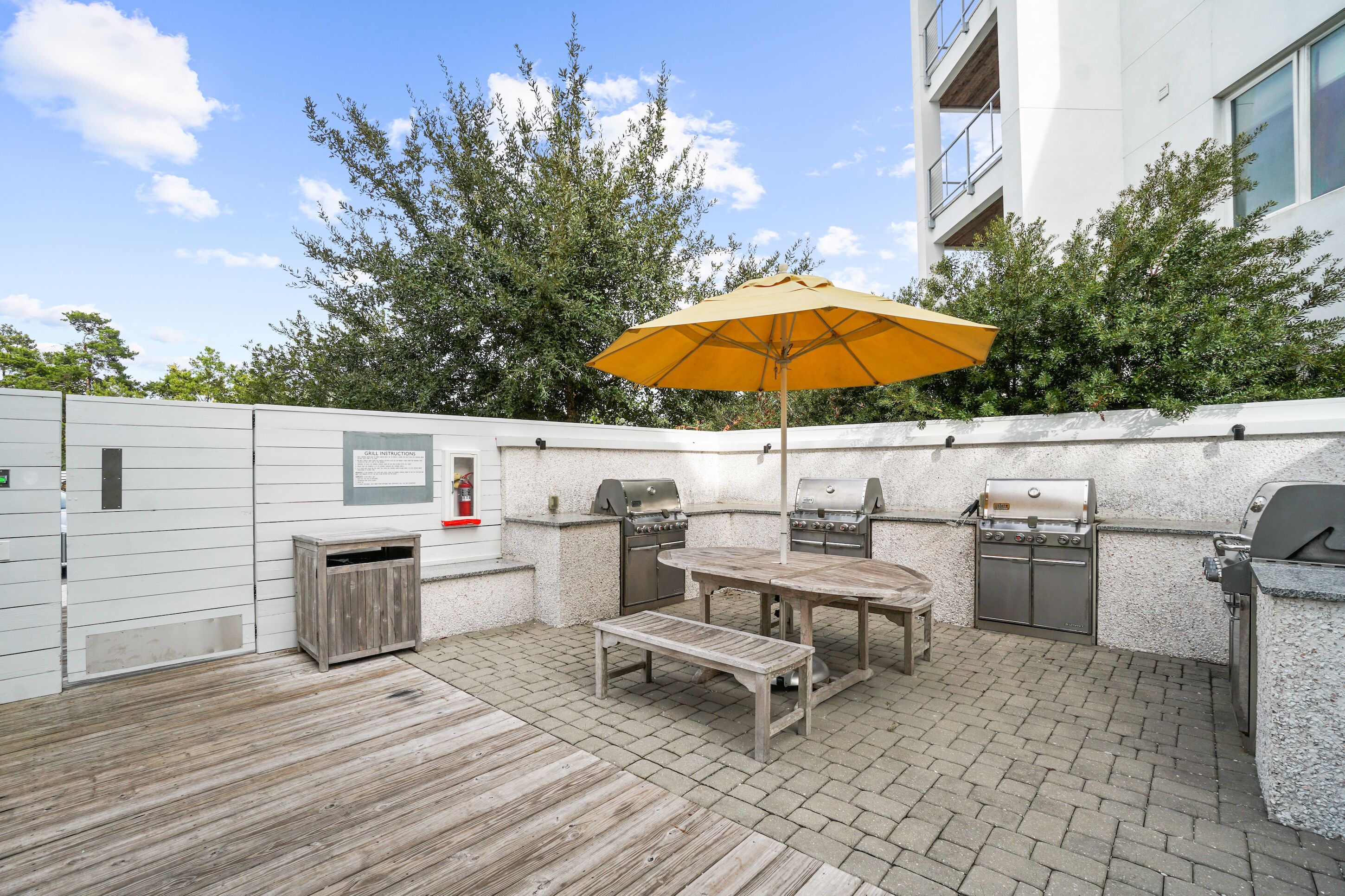 Un patio exterior con mesa y banco de madera, una sombrilla amarilla y una zona de barbacoa con extintor.
