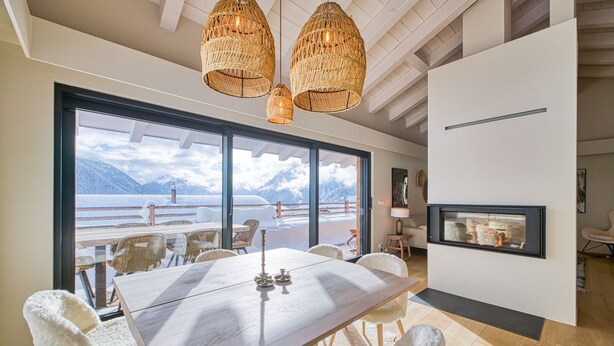 Uma sala de jantar moderna com uma mesa grande e cadeiras brancas, uma lareira e vista para montanhas através de grandes janelas.