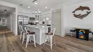 A modern kitchen with white cabinetry, a central island with bar stools, stainless steel appliances, and a decorative wall piece. - Villas at Seagrove by Panhandle Getaways (Santa Rosa Beach)