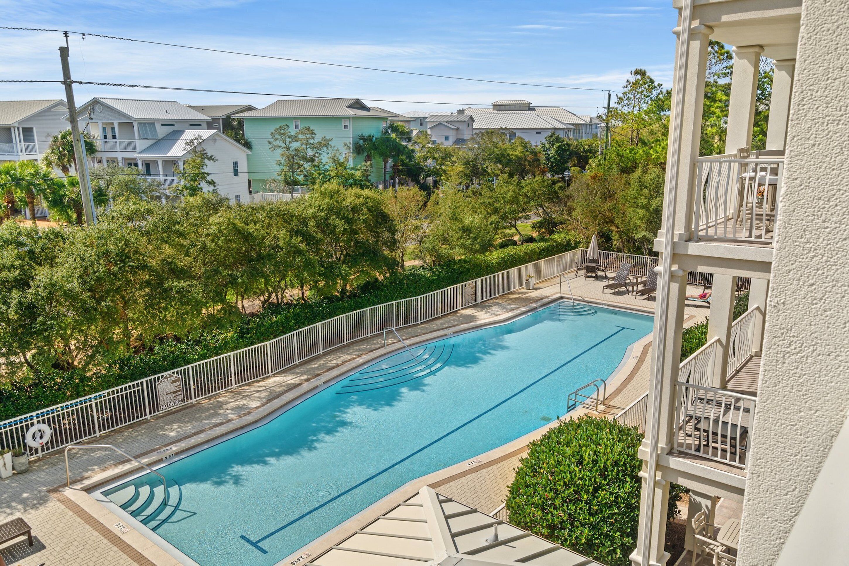 Piscina retangular com deck branco e grade de metal, rodeada por vegetação e edifícios residenciais.