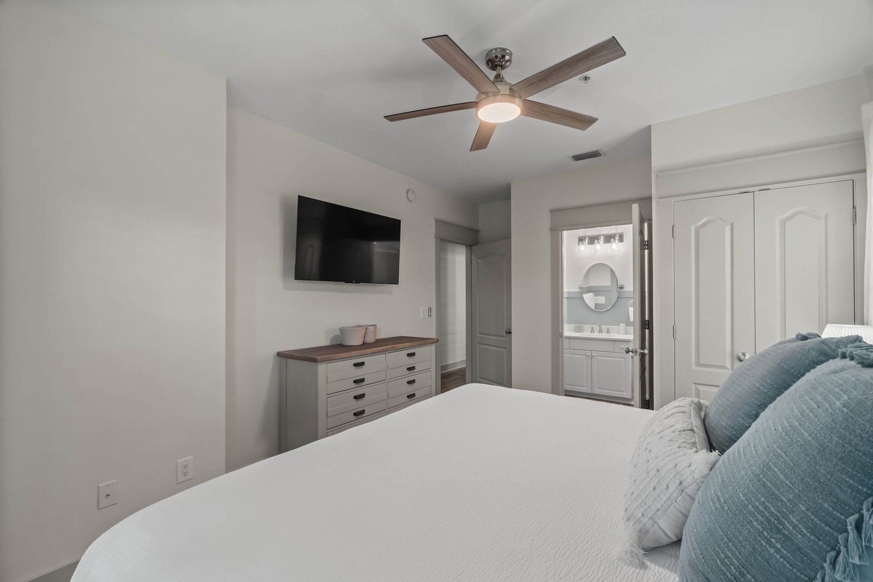 A bedroom with a bed, a ceiling fan, a TV, a dresser, and a door leading to a bathroom.