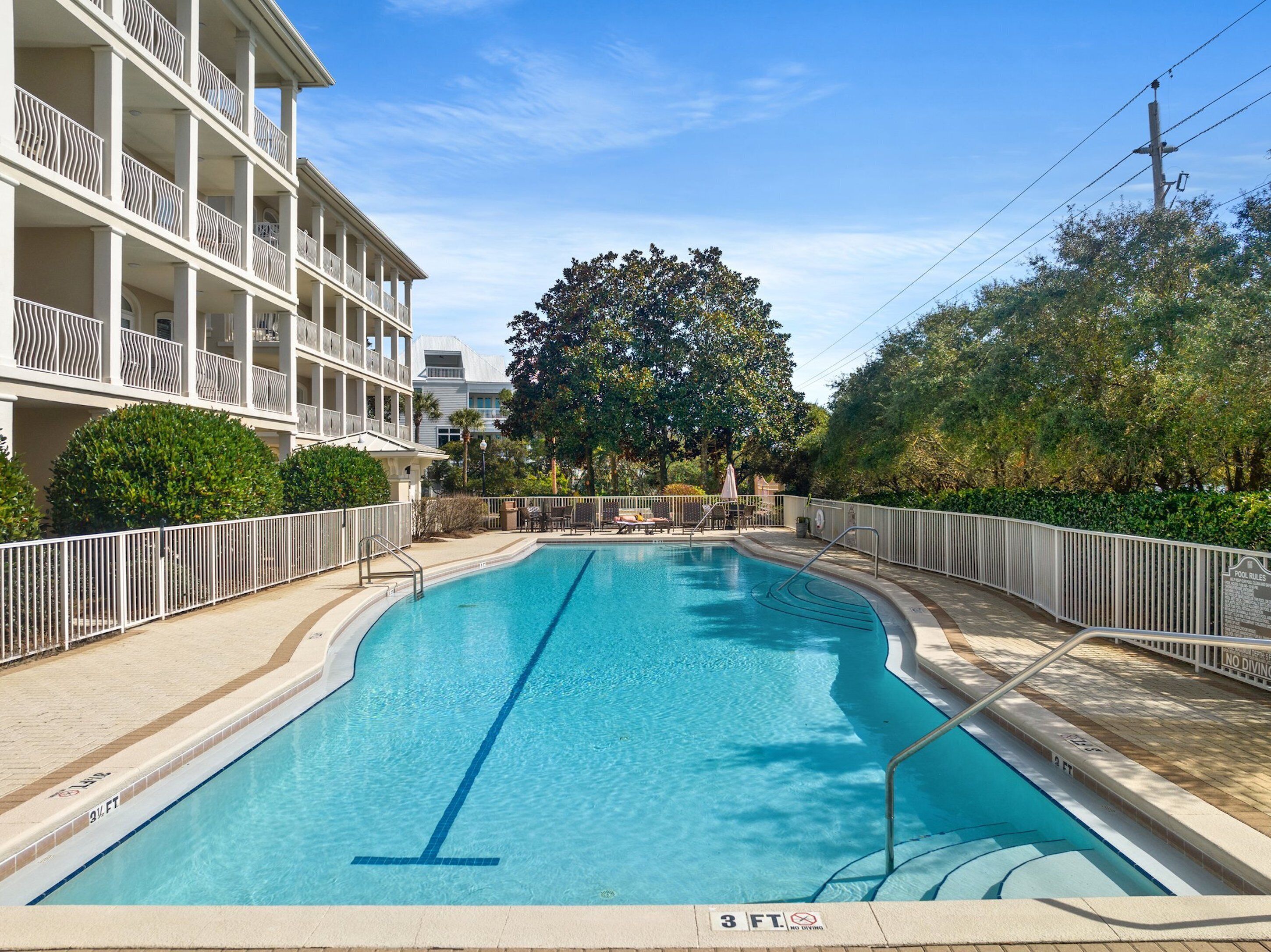 A swimming pool with a no diving sign, surrounded by a white fence, and adjacent to a multi-story building with balconies.