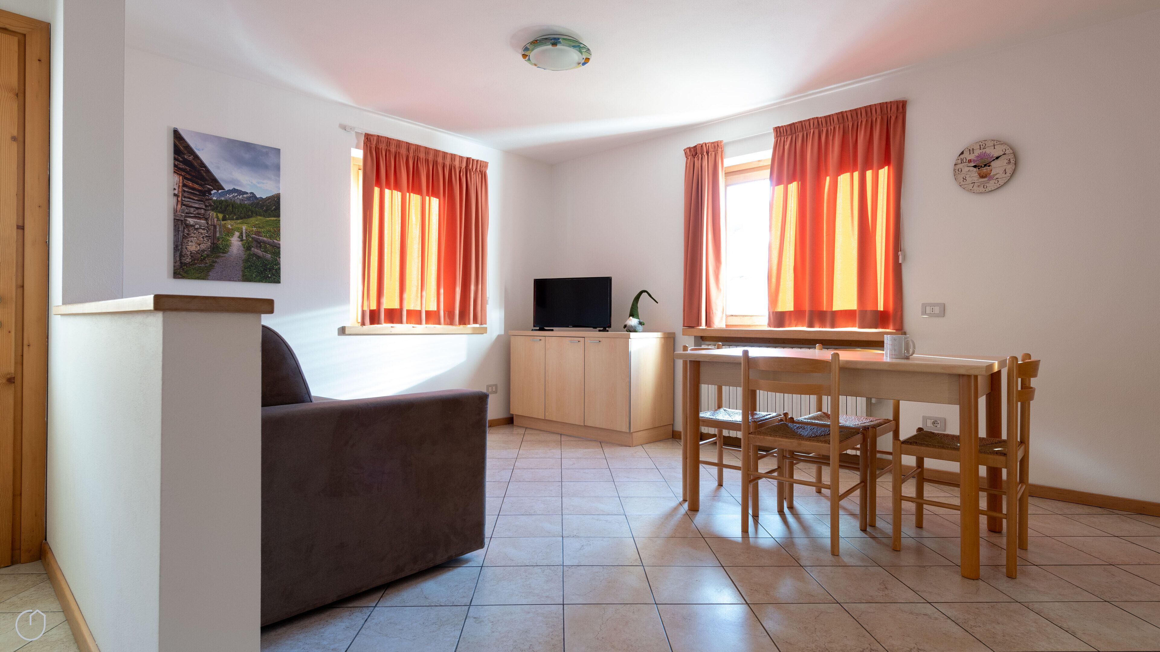 A small, well-lit room with a sofa, a TV, a dining table with chairs, and two windows with curtains.