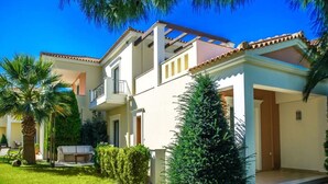 A two-story residential building with a balcony, a covered patio, and a well-maintained garden with palm trees and shrubs.