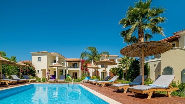 A swimming pool with lounge chairs and umbrellas, surrounded by buildings with a Mediterranean architectural style.