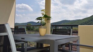 Un balcón con una mesa y sillas, una planta en maceta y vistas a colinas verdes bajo un cielo parcialmente nublado.