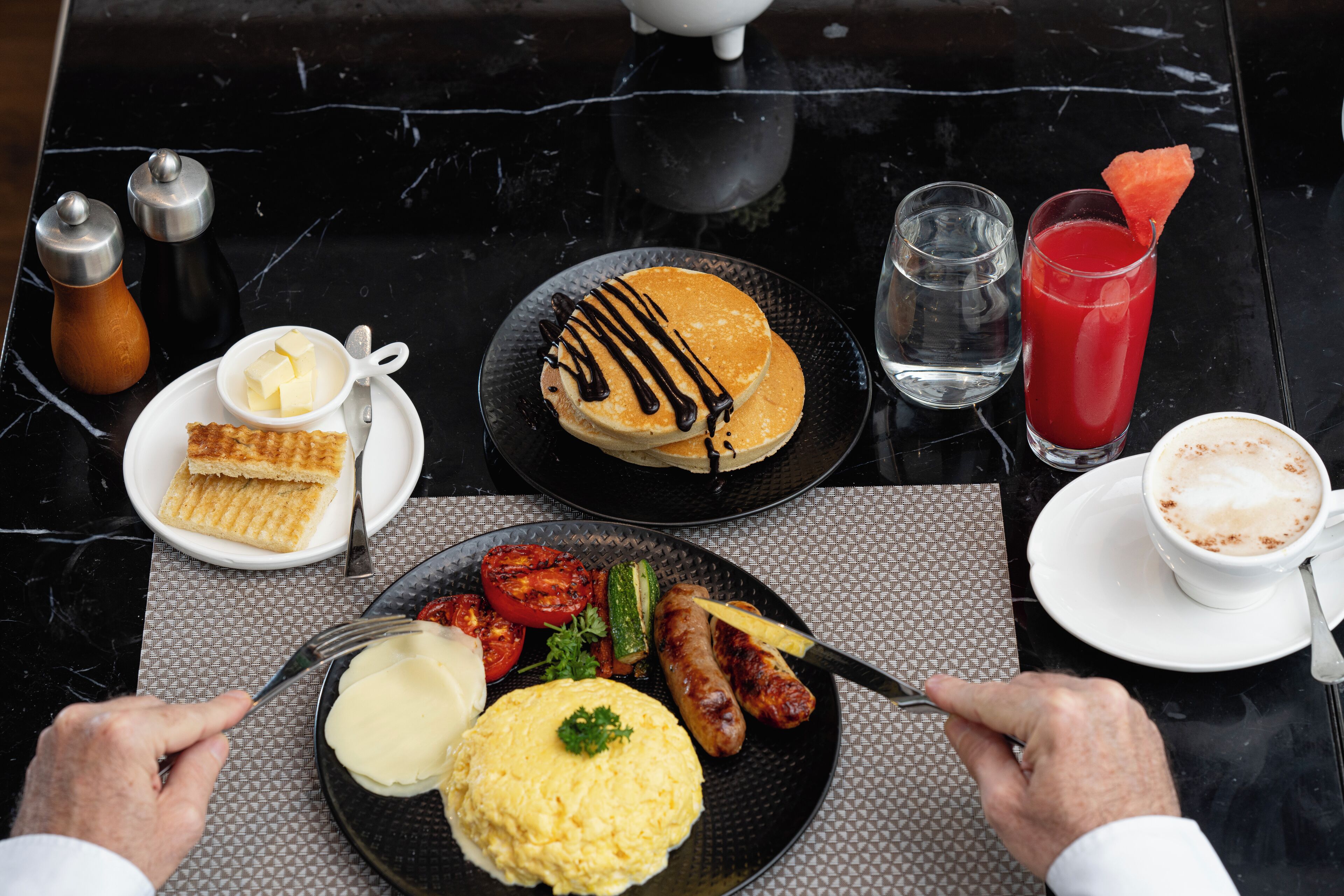 A breakfast setting with pancakes, scrambled eggs, sausages, and a red drink on a marble table.