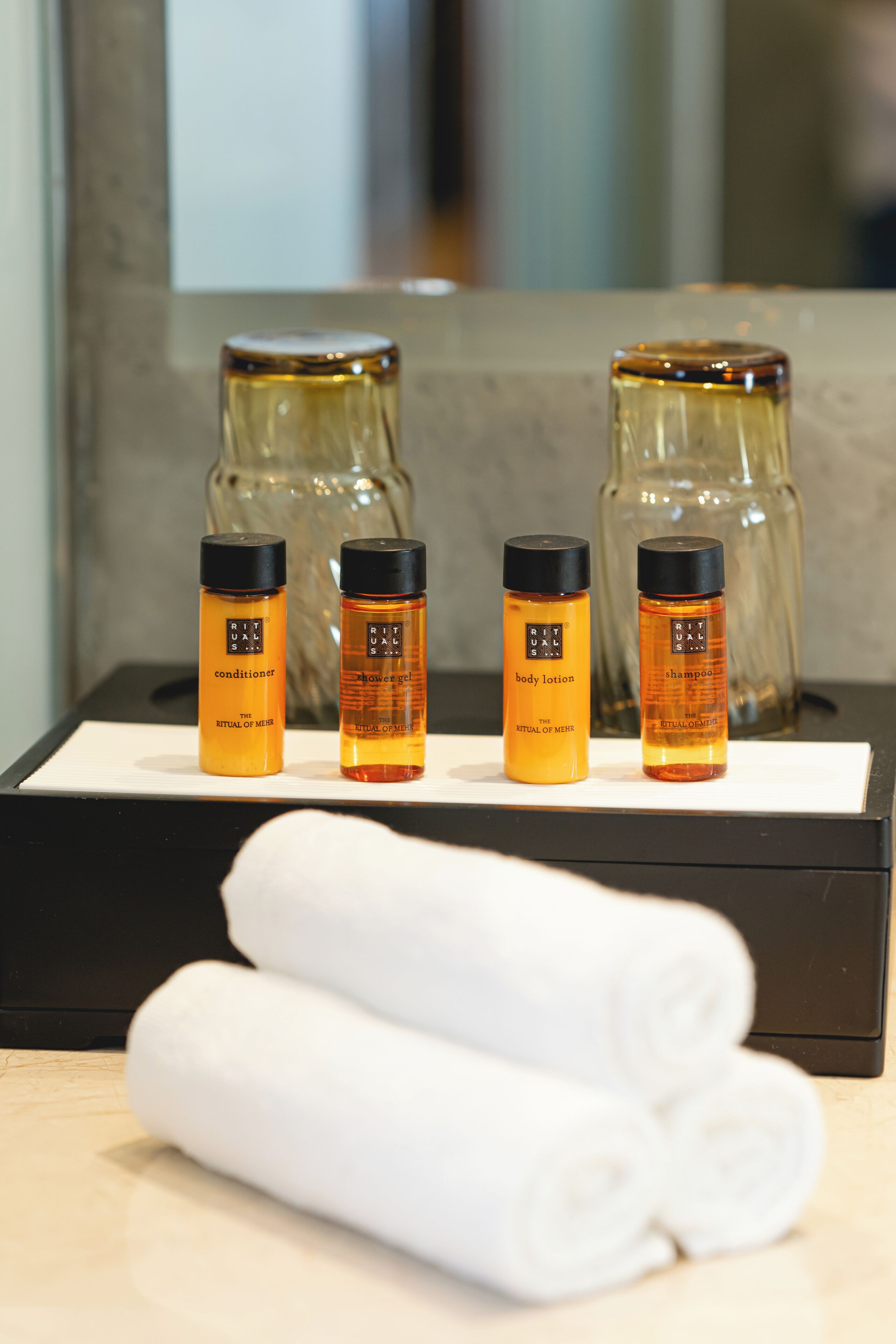 A hotel room with a black tray holding three bottles of Rituals body lotion, a bottle of shower gel, and a bottle of shampoo. There are two rolled white towels on the tray.