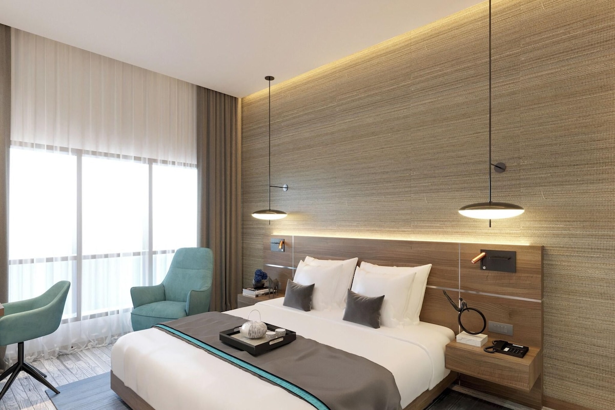 A modern hotel room with a large bed, two bedside tables, a nightstand, a desk, a chair, and a large window with curtains.