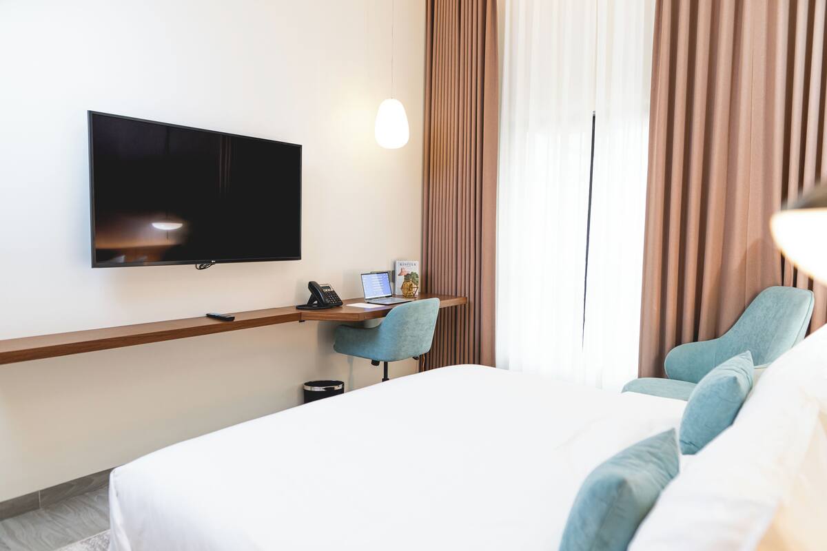 Una habitación de hotel con una cama, un escritorio con computadora, teléfono y televisor montado en la pared.