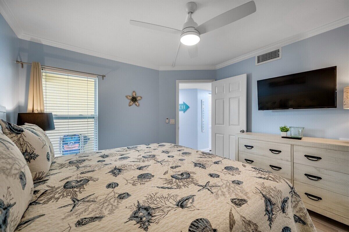 A bedroom with a bed, a ceiling fan, a TV, a dresser, and a door leading to another room.