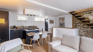 Ein modernes Wohnzimmer mit einer Couch, Sesseln und einem Essbereich mit Tisch und Stühlen. Es gibt eine Küchenzeile mit Herd und Spüle. Eine Treppe mit einer Steinwand ist sichtbar.