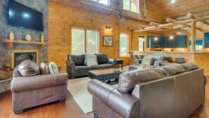 A log cabin interior with a stone fireplace, leather sofas, a flat-screen TV, and a kitchen area. - Sensational Views -4BR/New Hot Tub/New Firepit/Perfect for Families/No Event Fee (Gatlinburg)