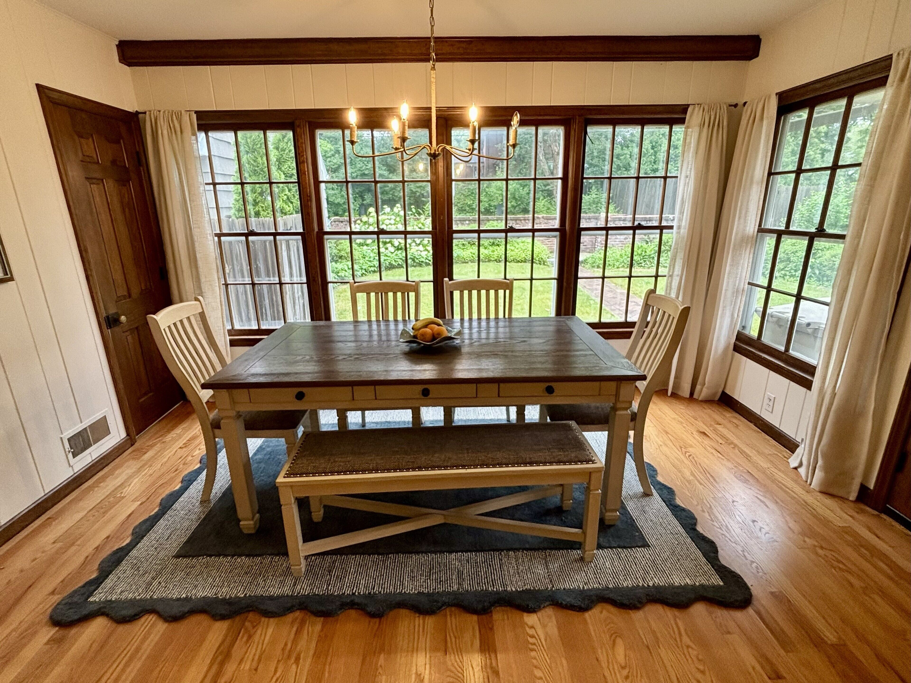 A dining room with a wooden table, four chairs, a chandelier, and large windows with curtains.